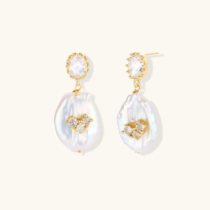 Gold-plated drop earrings with iridescent baroque pearls and crystal flower accents on cream background