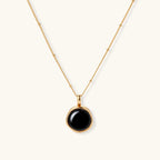 Circular black agate pendant on gold-plated sterling silver chain against cream background, showing modern minimalist design.
