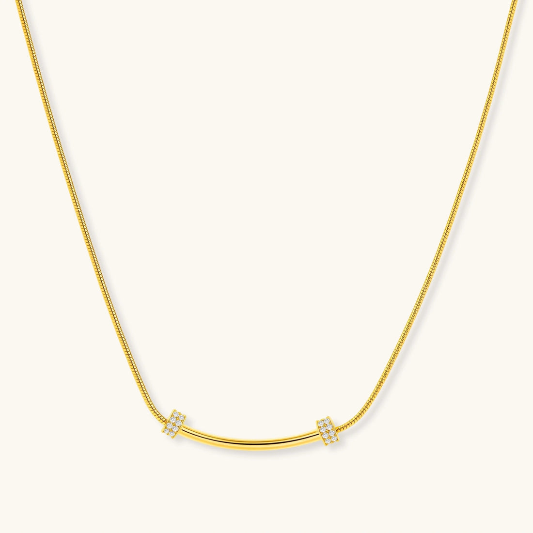 Delicate gold-plated necklace with curved bar pendant and crystal accents on snake chain against cream background.
