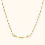 Delicate gold-plated necklace with curved bar pendant and crystal accents on snake chain against cream background.